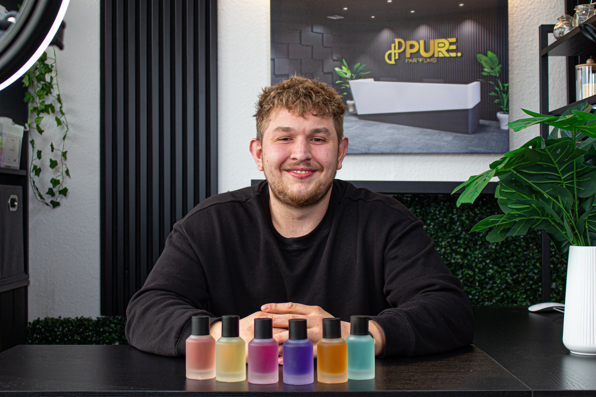 Nico, Mitgründer von Pure Parfums, sitzt stolz vor einer Auswahl der sechs farbenfrohen Parfümflaschen seiner Marke. Die leuchtenden Farben der Flaschen heben sich gegen den neutralen Hintergrund ab und symbolisieren die Vielfalt und Qualität der Düfte, die Pure Parfums bietet.