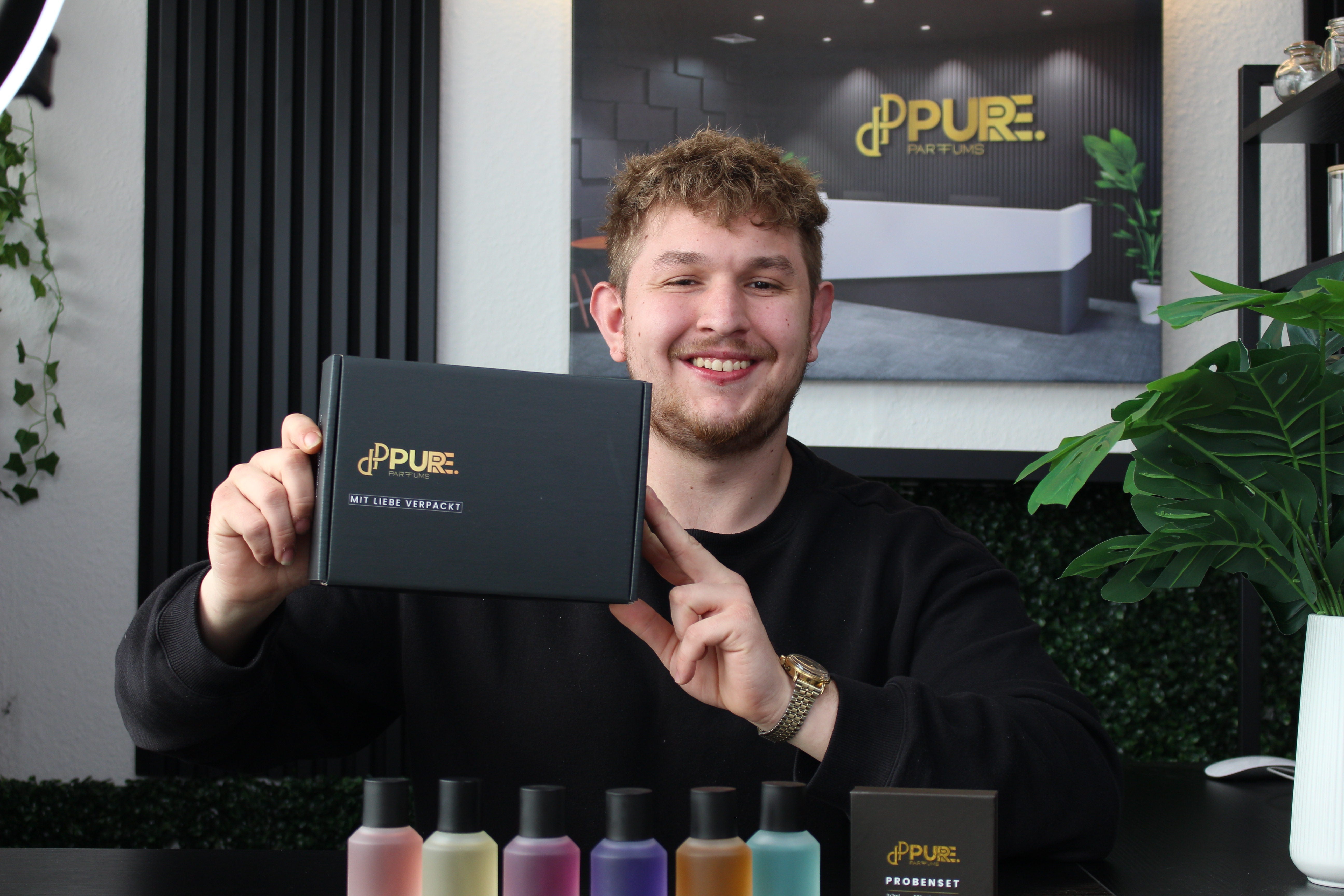 Nico, Mitgründer von Pure Parfums, präsentiert stolz einen Versandkarton mit einem einzigartigen, personalisierten Design, das die Markenidentität widerspiegelt. Er hält den stilvoll gestalteten Karton, der die Premium-Qualität und Detailgenauigkeit der Marke unterstreicht.